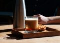 Gals mit schaumigem Nitro Coffee und Nusskeks auf Holztablet, dahinter ein Teil von einem Sahnespender und ein Teil von einem Arm