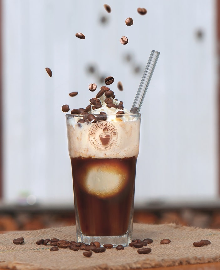 Glas mit Eiskaffee und Strohhalm, auf das von oben herunter Kaffeebohnen herabrieseln