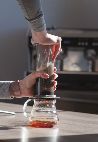 Frauenhand drueckt von oben herab Aeropress, Kaffee tropft unten heraus in eine Glaskanne