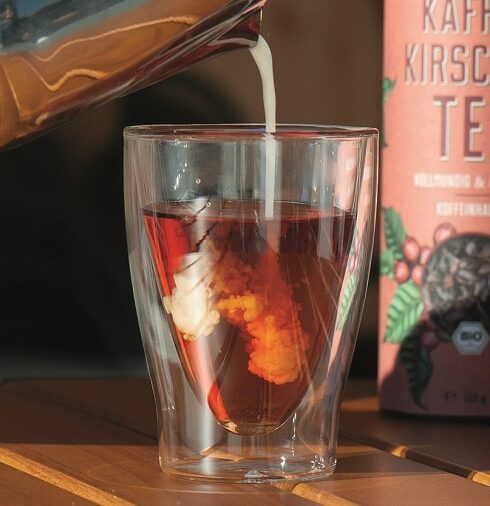 Glas mit durchsichtiger rotem Kaffeekirschentee Cascara in das von oben Milch eingegossen wird
