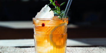 Glas mit gelber Fluessigkeit, Eiswuerfeln, Zitronenscheiben, Cranberrys und einem Strohhalm, von oben kommt eine Hand herab mit Rosmarinzweig Rezept für Cascara