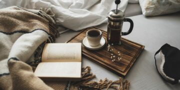 Coffee brewed in a French press and a cup on a wooden board with open book in bed.