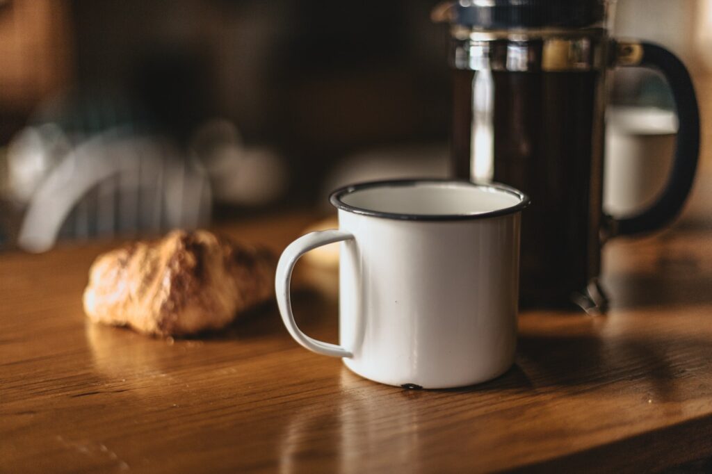 enamelled cup of coffee on wooden table in front of a croissant and a french press