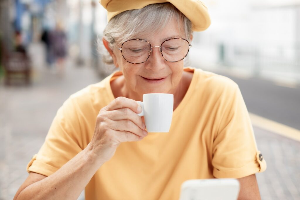Portrait of smiling attractive elderly woman in yellow enjoying an espresso coffee cup lookingphone