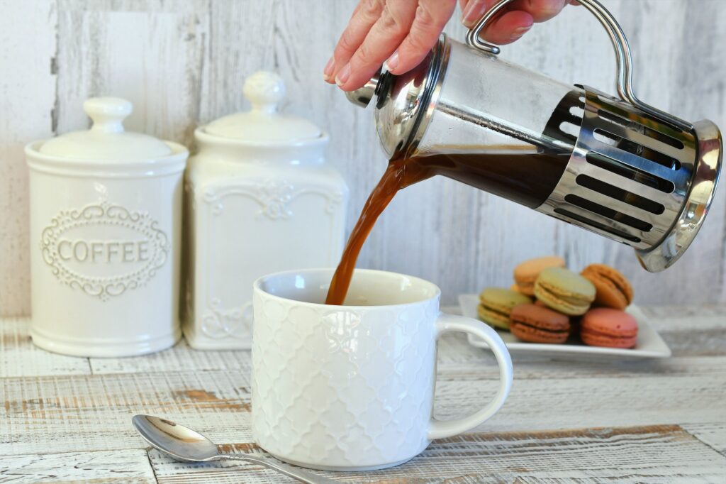 Pouring coffee from French press coffee maker into white mug with plate of French macaron cookies