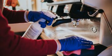 Woman cleaning the coffee machine with sanitizer and towel with blue gloves put on