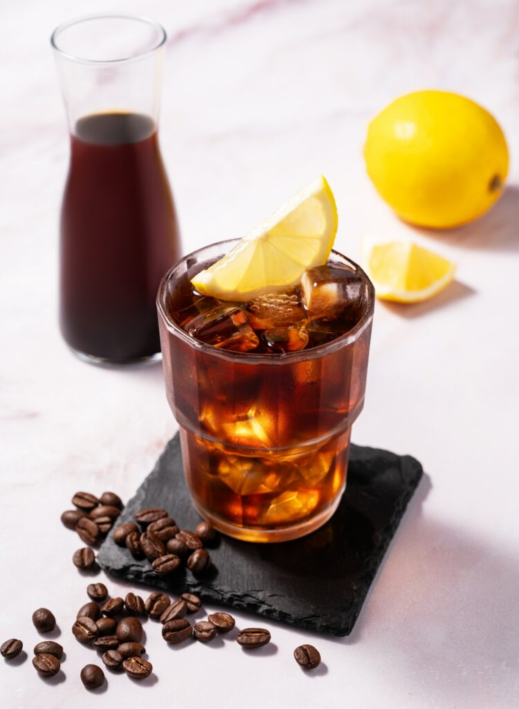 Cold brew coffee in a glass with ice and lemon on a light marble background with coffee beans