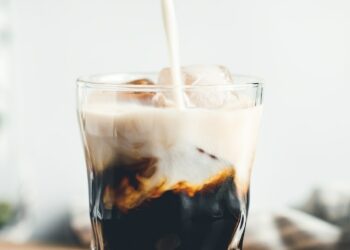 Pouring milk in a glass with espresso and iced cubes in a modern kitchen in the morning.