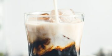 Pouring milk in a glass with espresso and iced cubes in a modern kitchen in the morning.