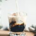 Pouring milk in a glass with espresso and iced cubes in a modern kitchen in the morning.