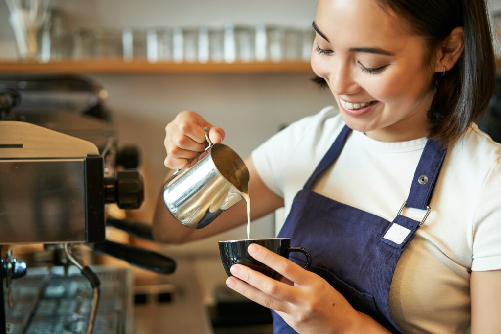Smiling asian barista girl, making coffee, pouring steamed milk into cappuccino, doing latte art in