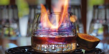 Glas auf gusseisernem Tablett mit etwas Rum und Kaffee befuellt, einer Feige daneben und einer Flamme im Glas
