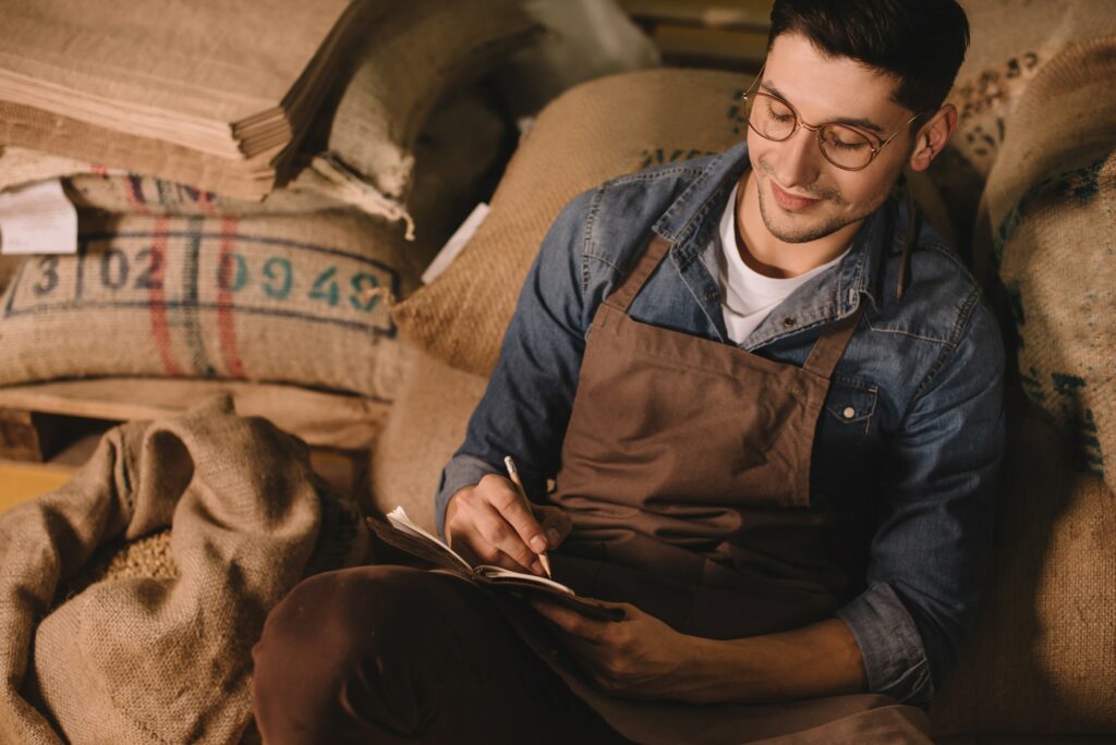 portrait of smiling coffee roaster in eyeglasses and apron making notes in notebook