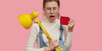 Indoor shot of depressed sad woman opens mouth, cries desperately, holds red cup of tea, desk lamp,