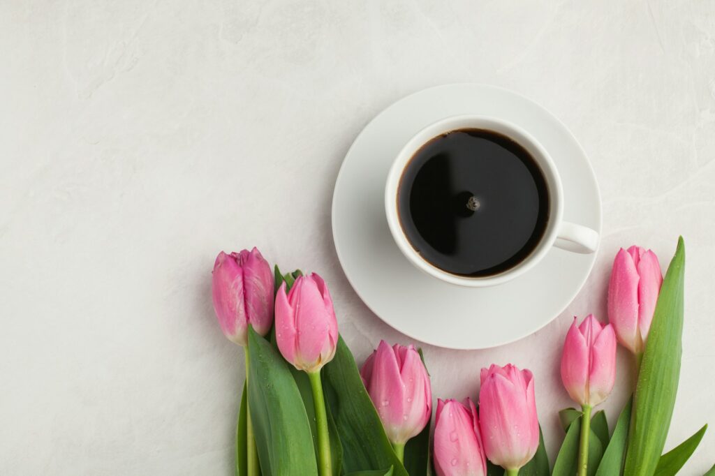 Black coffee in white Cup with pink tulips on light stone background. Top view with copy space